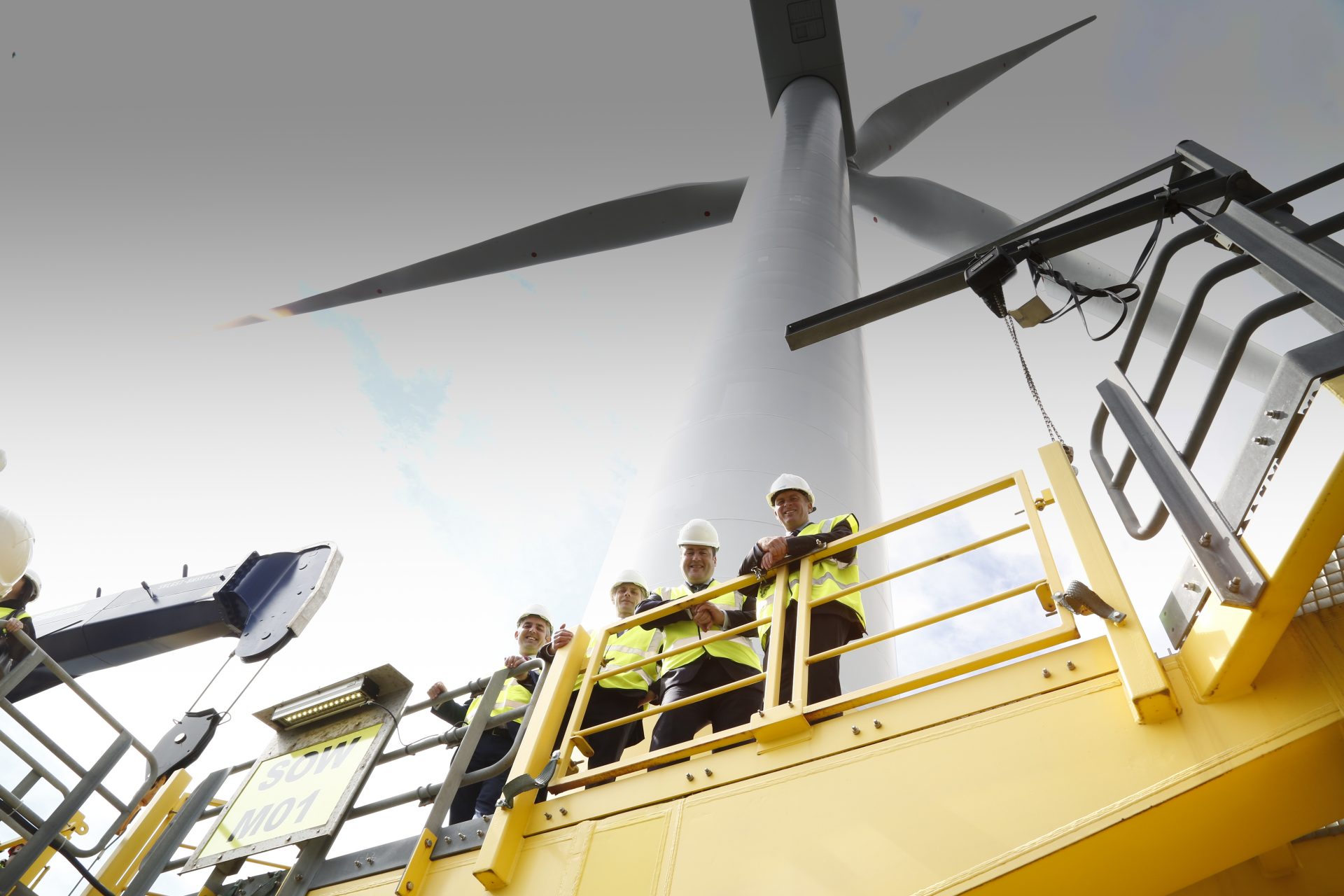 Unos obreros se sitúan en la plataforma base de un aerogenerador y miran hacia abajo, la turbina eólica se vislumbra al fondo.
