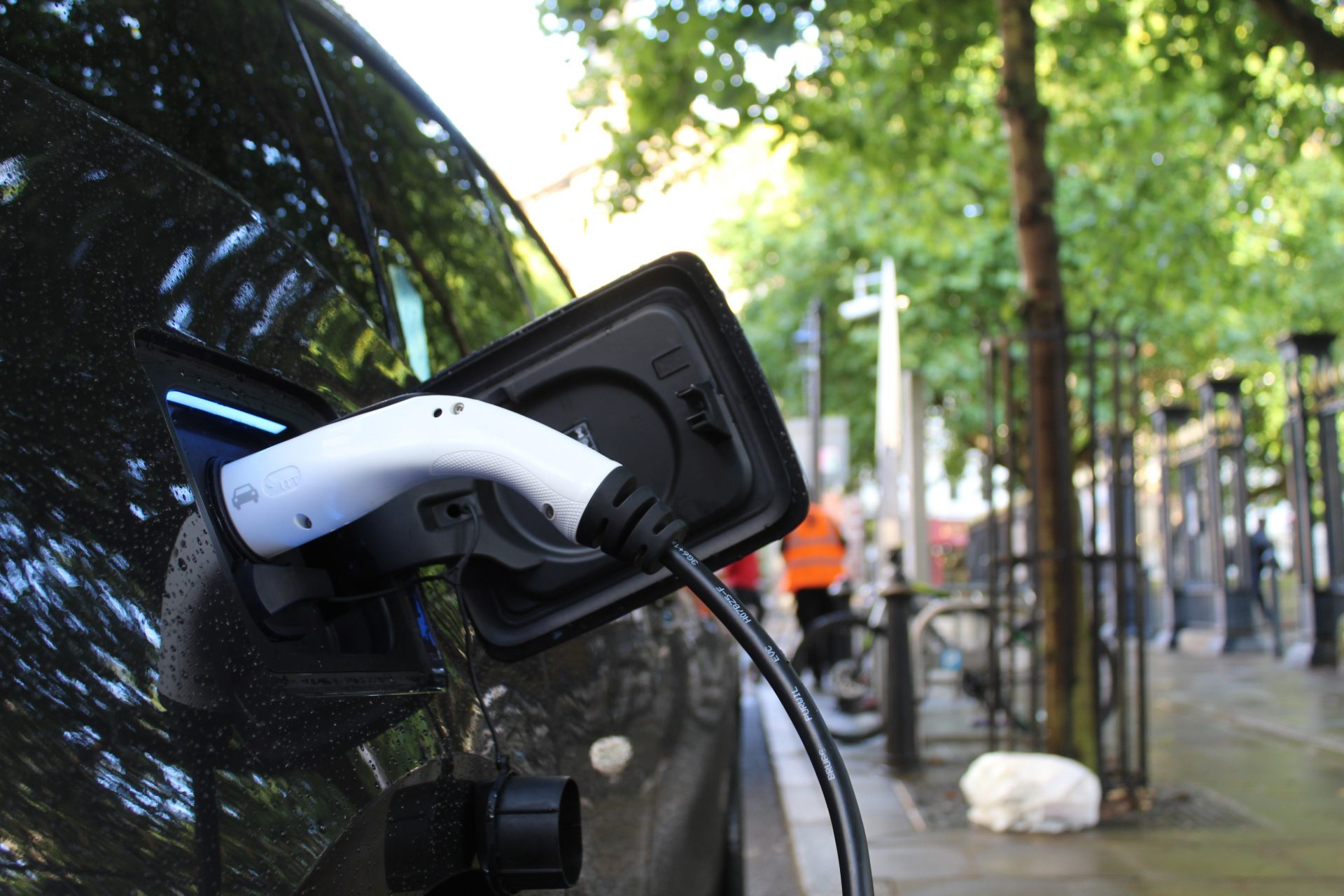 Ein Ladekabel steckt in einem E-Auto, das am Straßenrand parkt. Im Hintergrund grüne Bäume.