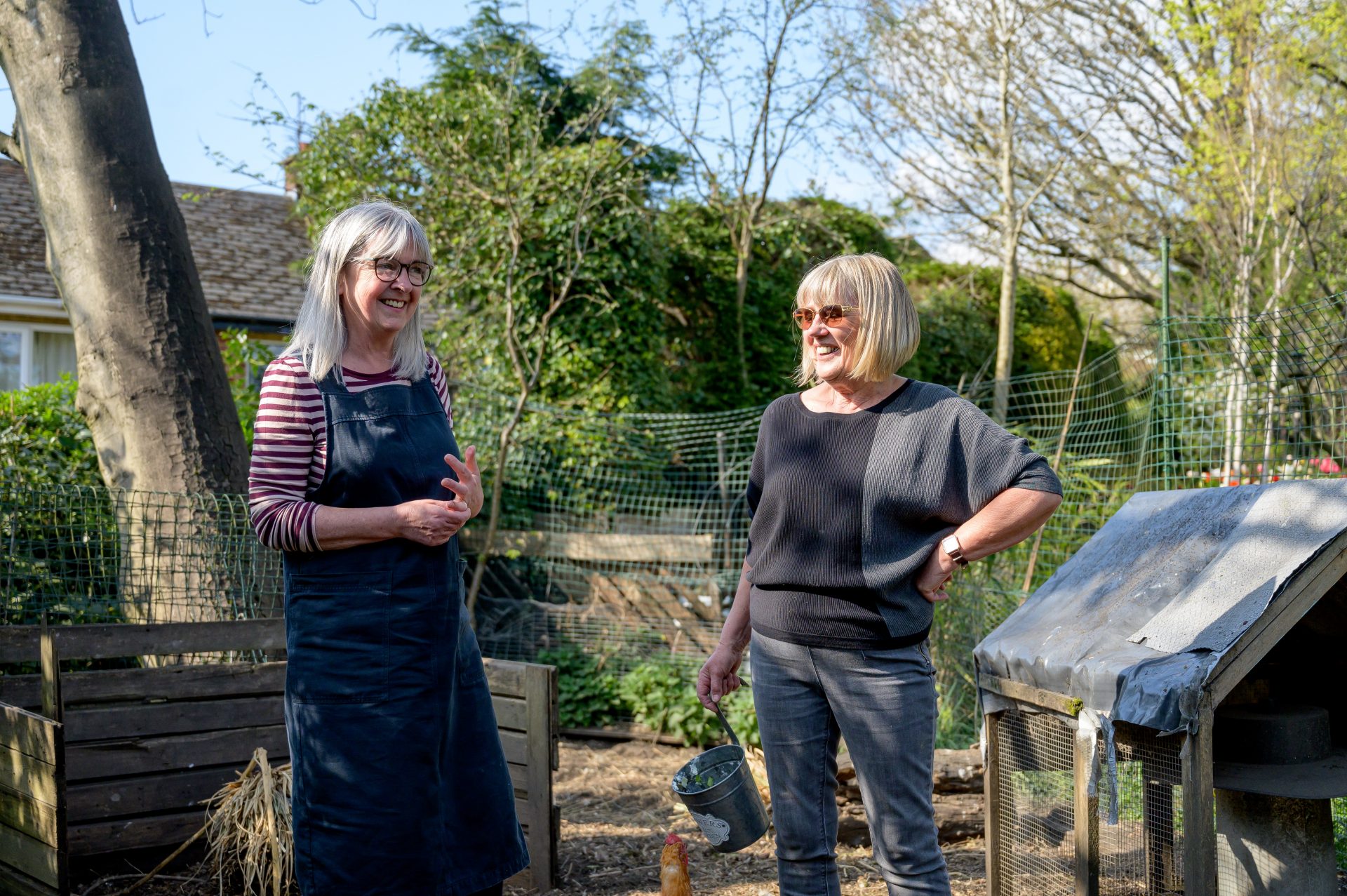Zwei ältere Frauen stehen in einem Garten in der Sonne und unterhalten sich lachend.