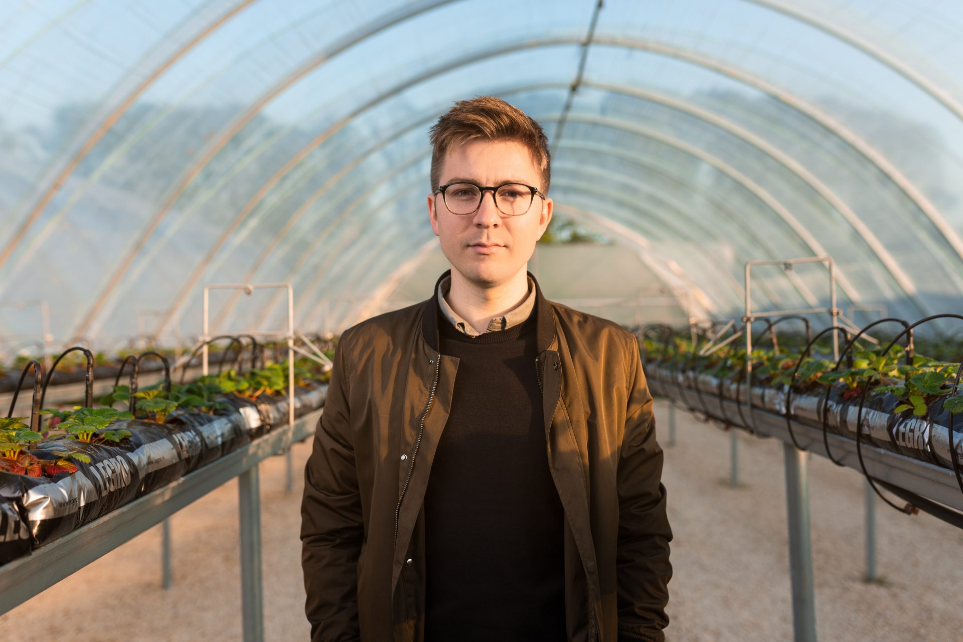 Ein junger Mann steht in einem Gewächshaus, im Hintergrund sind Tische mit Jungpflanzen aufgereiht, die mit Bewässerungsschläuchen versehen sind.