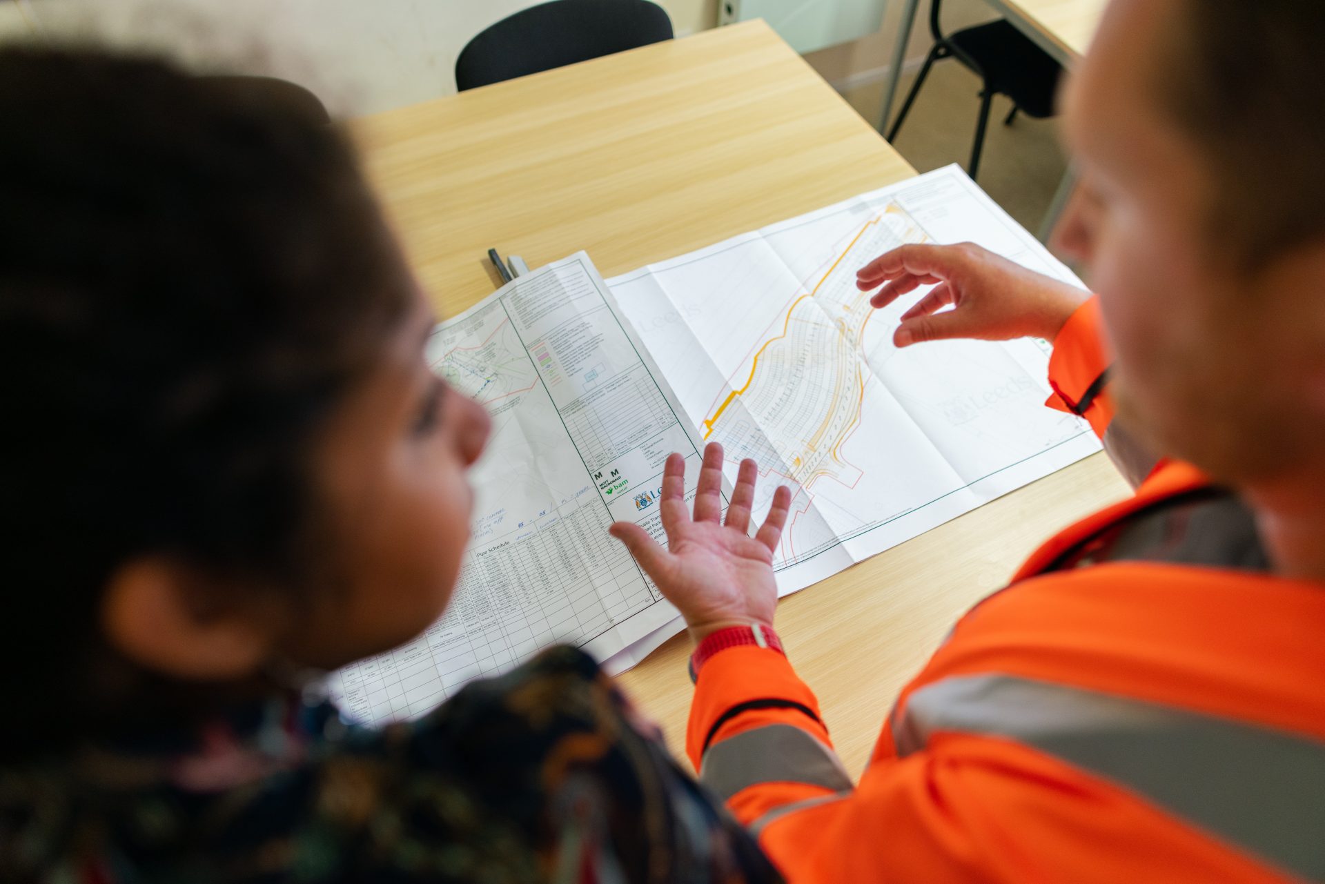 Zwei Personen stehen vor einer Planungskarte, die auf dem Tisch liegt. Eine der Personen trägt eine orangene Warnweste.